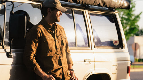 A man wearing a brown overshirt, blue jeans, and a cap leans against the side of a vintage beige SUV with a roof tent, in an outdoor street setting during golden hour.